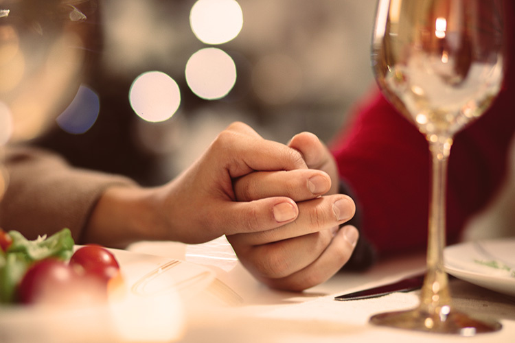 Mies ja nainen pitävät toinen toistaan kädestä kiinni ravintolan pöydän ääressä. Pöydällä myös viinilasi.