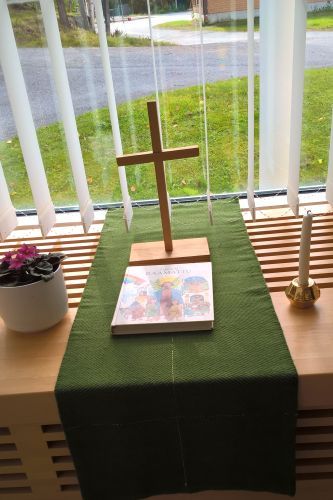 Ikkunan edessä pöydällä puinen risti, lastenkirja, kynttillä ja ruukkukukka.