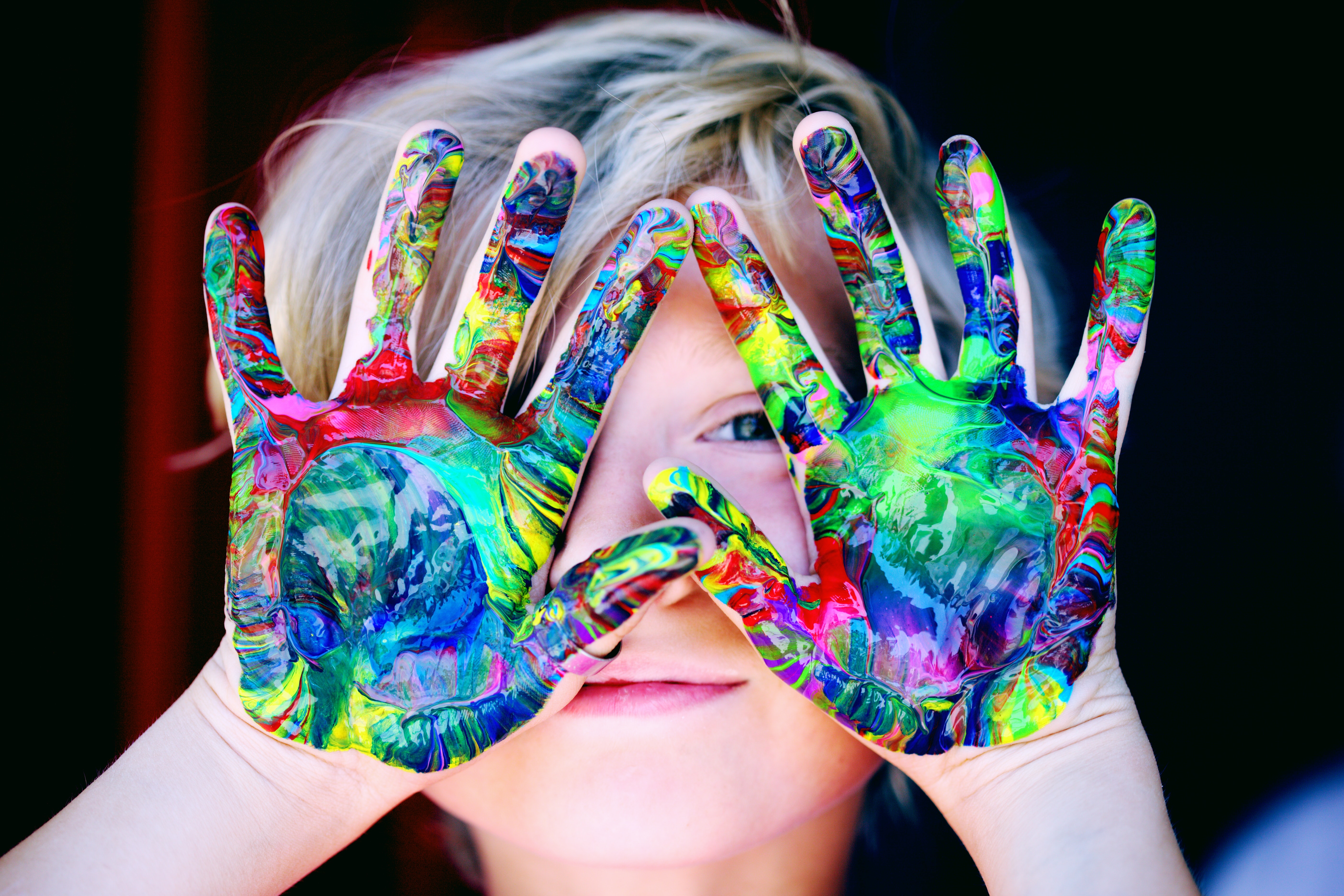 Lapsi kurkistaa värikkäiksi maalattujen käsiensä takaa.