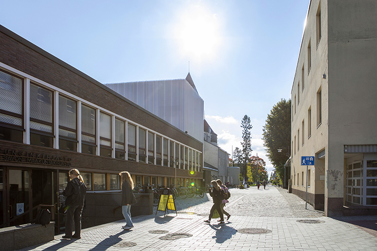 Vasemmalla ihmisiä menossa Kuopion pääkirjastoon, oikealla Postitalon kuva. Aurinkoinen kesäpäivä.