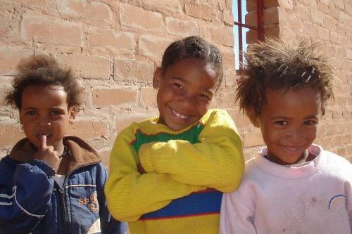 Kolme Kalaharin lasta. Yksi pitää sormea suun edessä ja kaksi muuta hymyilee.