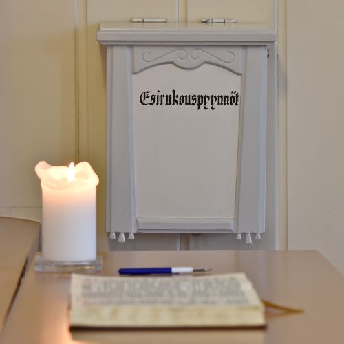 Etualalla avoin Raamattu ja palava kynttilä, taustalla Tuomiokirkon harmaansävyinen esirukouspyyntölaatikko.