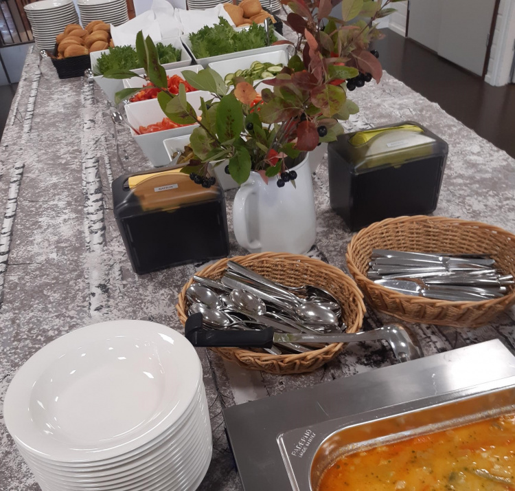 Pöydässä syviä lautasia, keittoastia, kauha, lusikoita, veitsiä seviettejä, kukkia tomaattia sämpylöitä, salaatinlehtiä kupissa