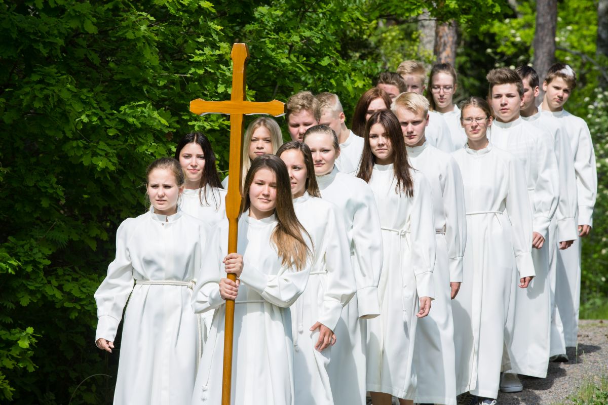 Konfirmaatiomessu / Hirvijärvi 1 Konfirmoitavia nuoria kävelemässä jonossa kirkkoon albat päällään. Etummaisella nuorella on puuristi kädessään.