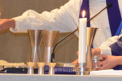 Pappi asettamassa ehtoollista. Pöydällä ehtoollistarvikkeita ja palava valkoinen kynttilä.