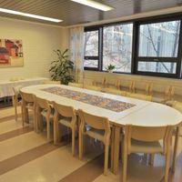 Kerhohuone Aaron Ovaalinmutoinen pöytä ja tuolit ympärillä. Ikkuna taustalla.