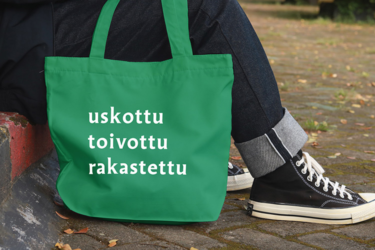 Henkilö istuu ulkona kiviportailla, jalkojen vieressä kangaskassi, jossa teksti uskottu toivottu rakastettu